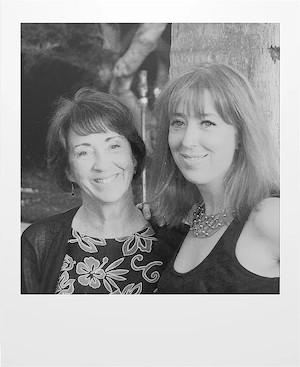 Two women stand close together and smile at the camera. One has short dark hair and wears a floral top, while the other has long light hair and wears a sleeveless top with a necklace. Trees are visible in the background.