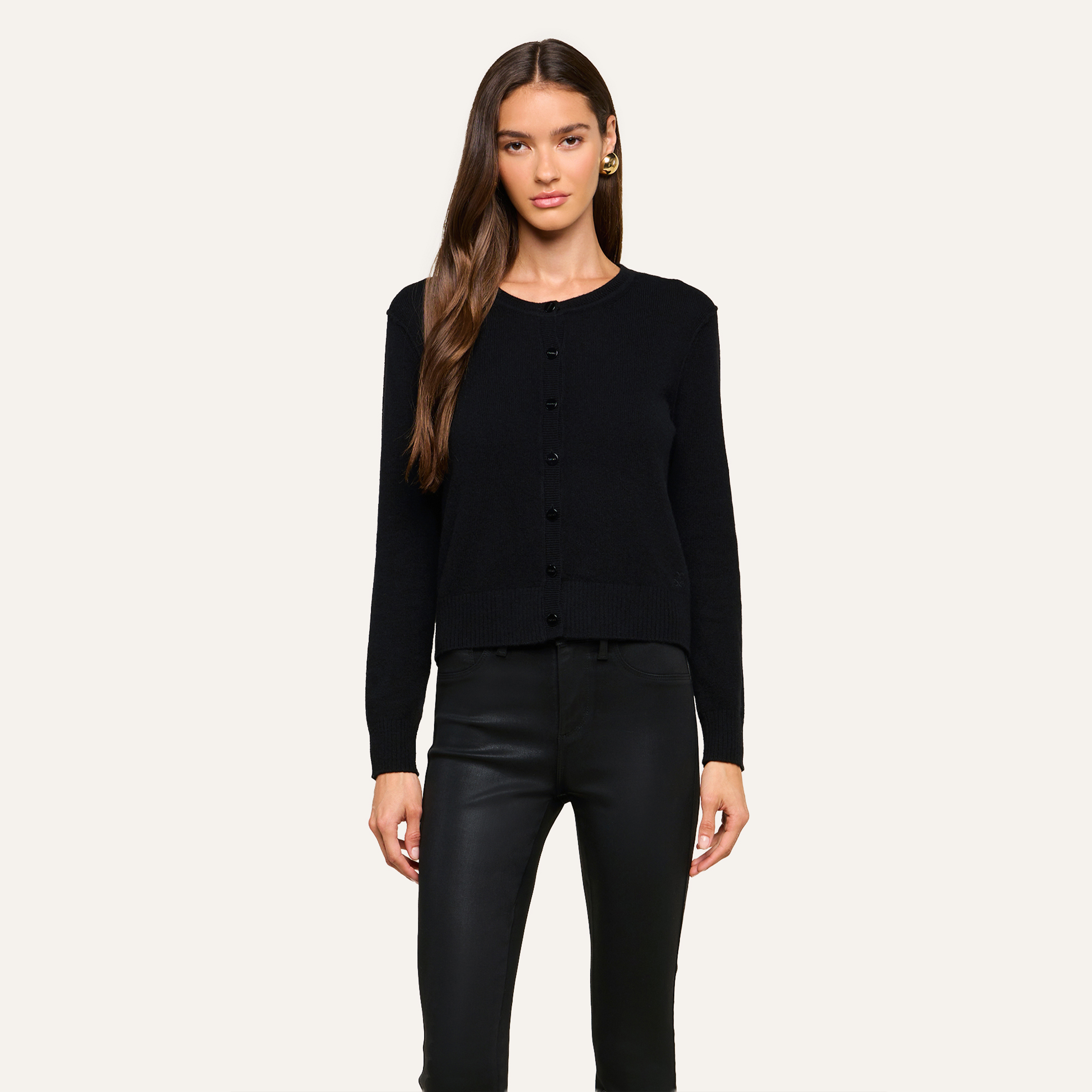 Woman standing against a plain background, dressed in a black button-up cardigan and black leather pants, with long brown hair and gold hoop earrings for a sleek, modern look.