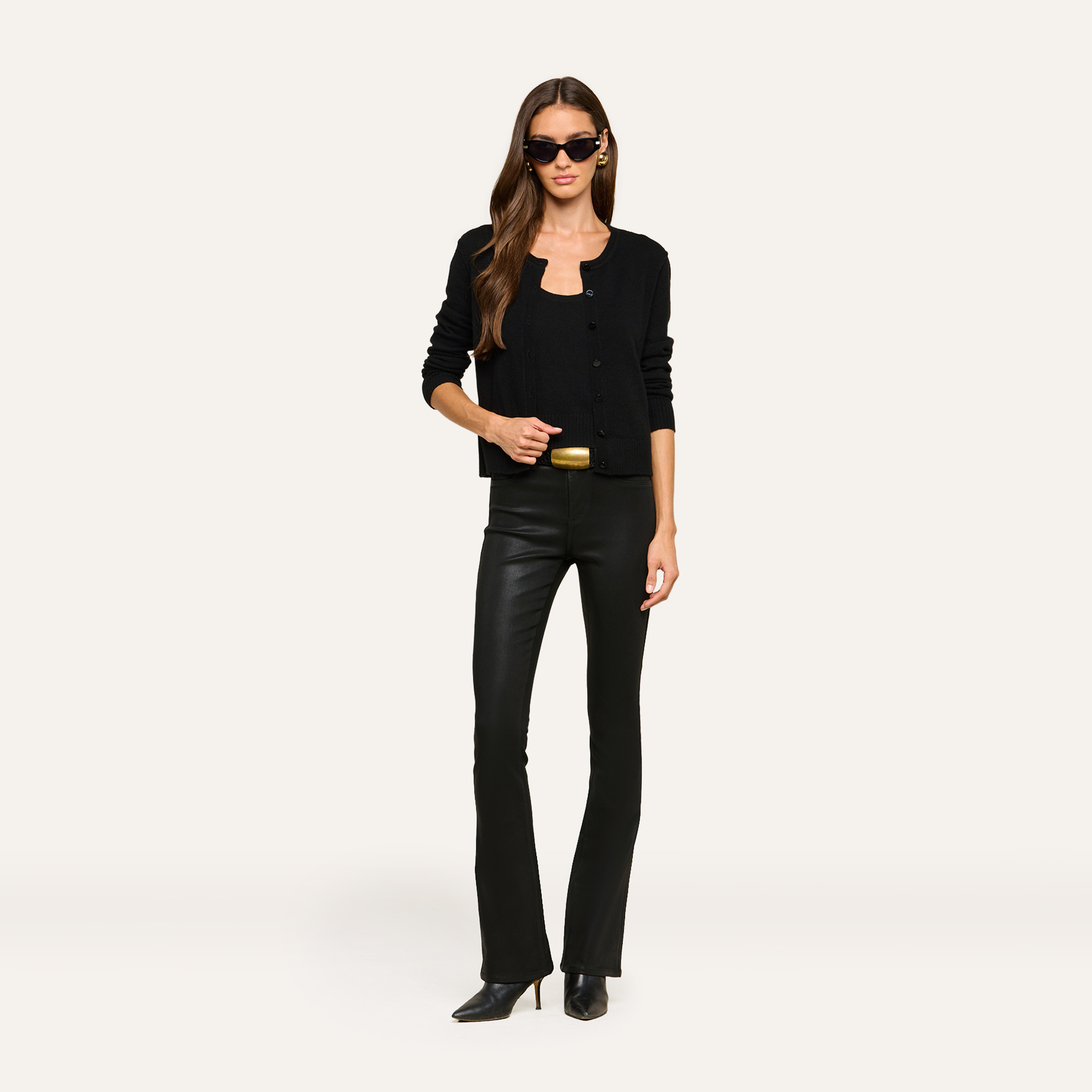 A woman wearing black sunglasses, a black cardigan, black leather pants, and black heeled boots stands against a plain light background, holding a gold clutch at her waist. Her chic style is accentuated by the sleek leather pants.