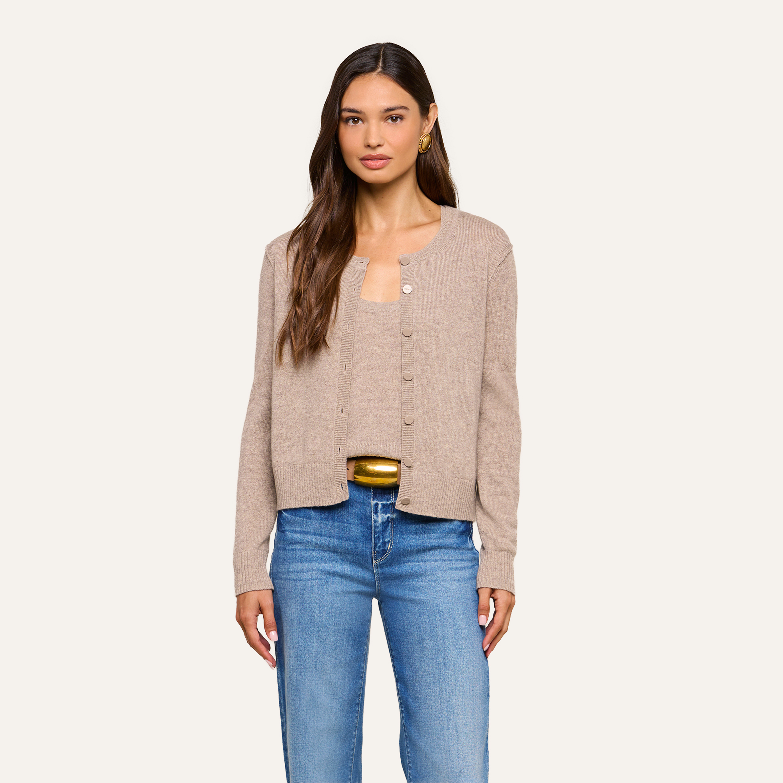 Woman with long brown hair wearing a beige cardigan over a matching top and blue jeans, standing against a plain light background, looking forward with a neutral expression.