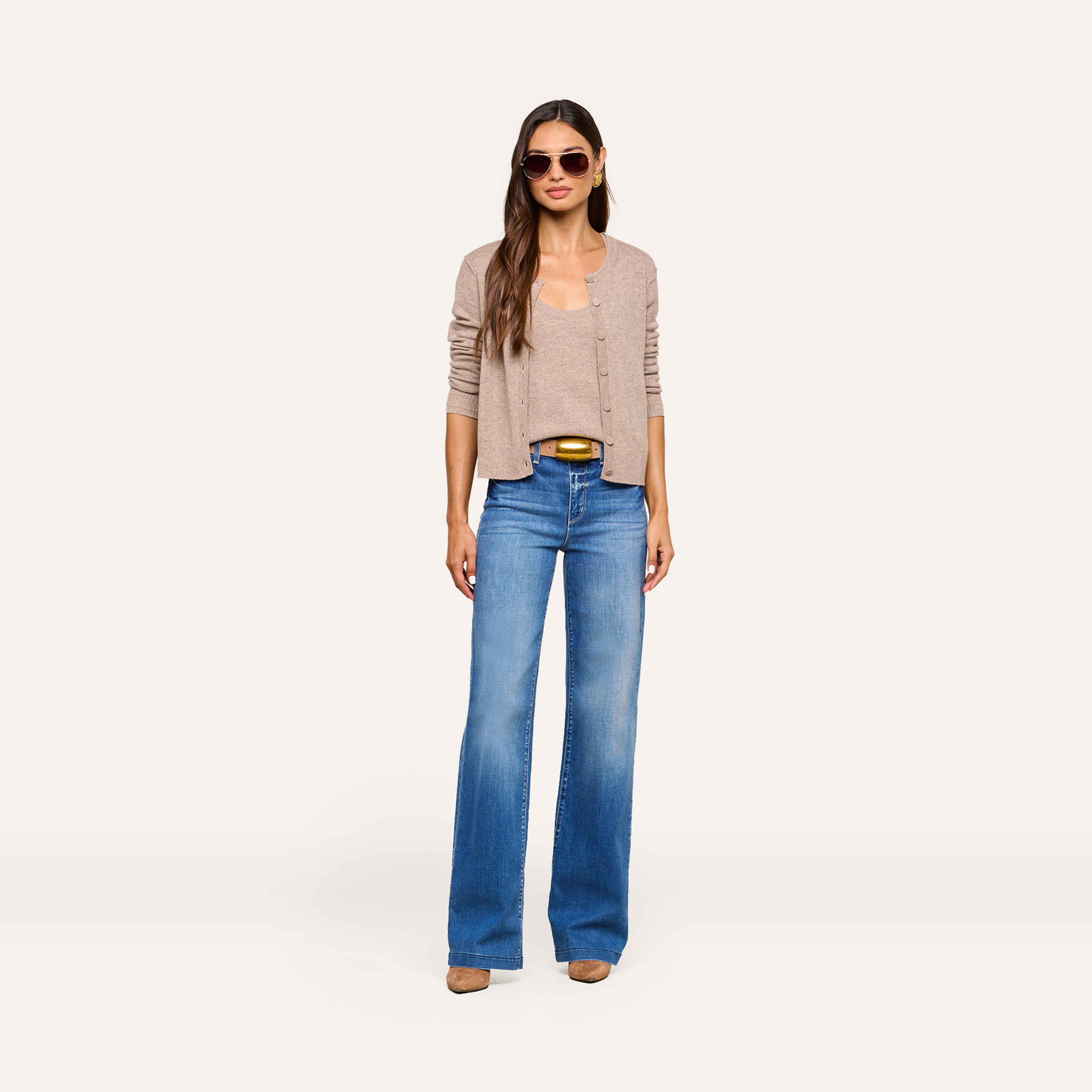 A woman wearing sunglasses, a beige cardigan over a matching top, blue wide-leg jeans, and brown shoes stands against a plain light background, exuding effortless style.