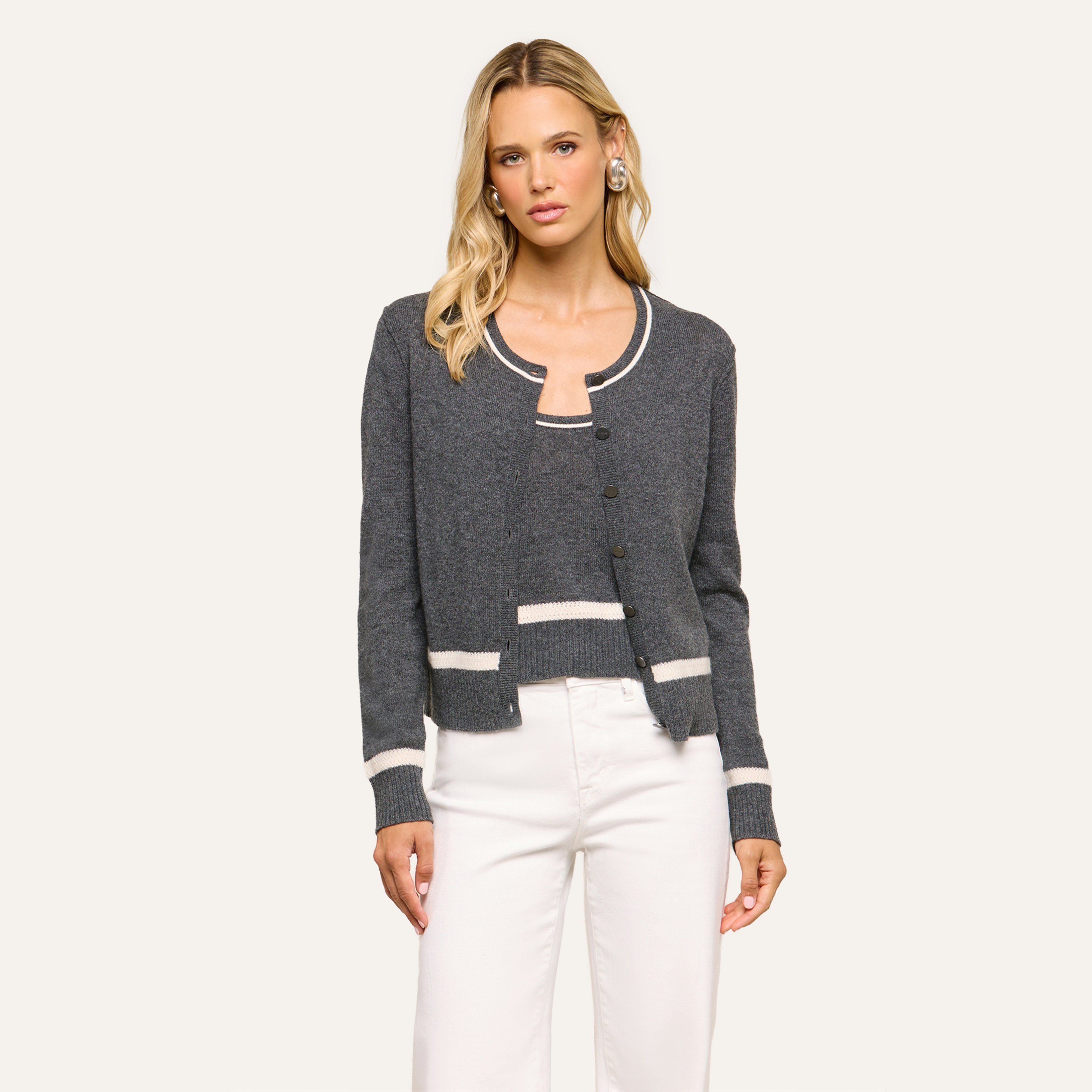 Woman wearing a charcoal gray cardigan with white trim over a matching top and white pants, styled in an elegant look, standing against a plain light background and looking at the camera.