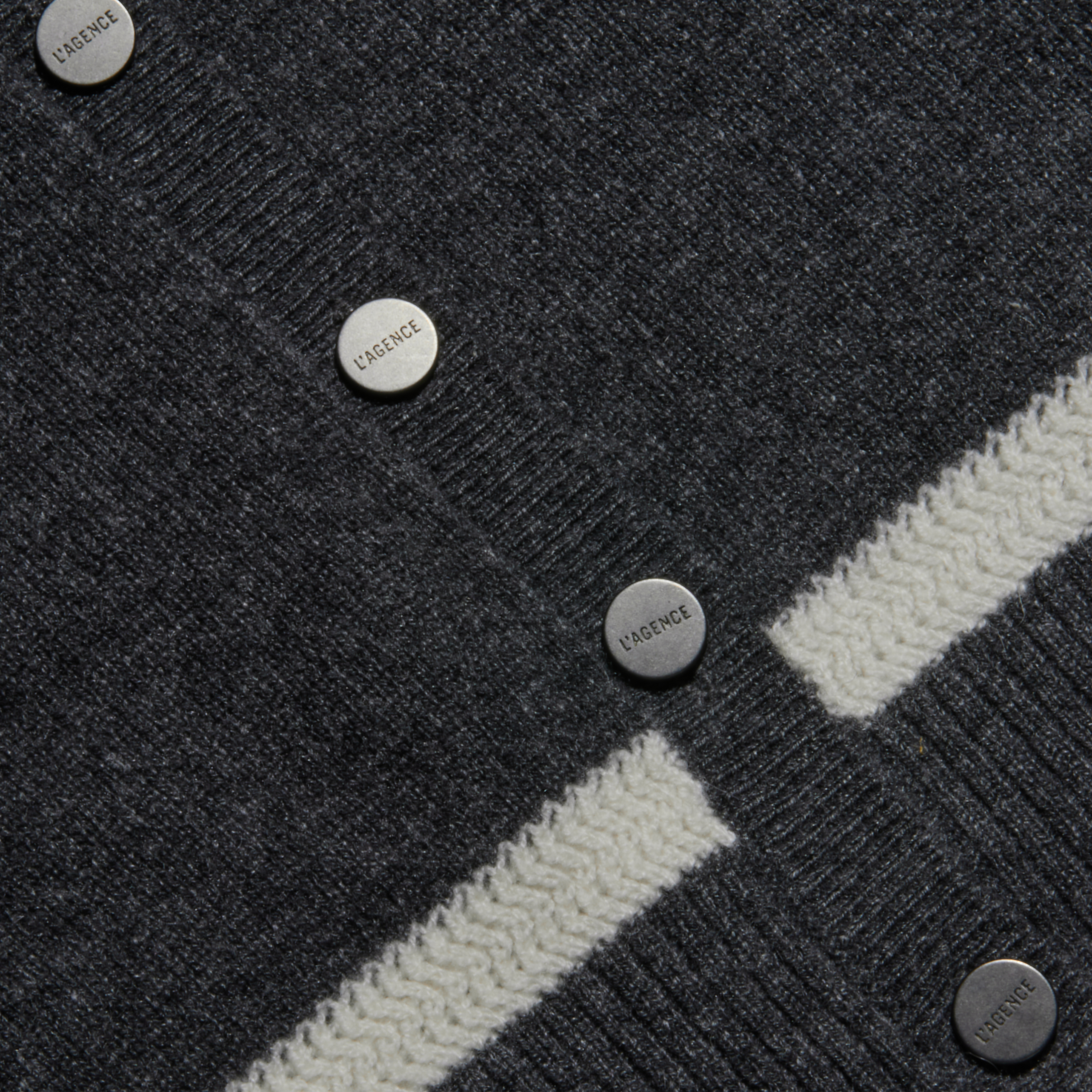 Close-up of a dark gray knit fabric with metal buttons labeled LAGENCE, featuring a cream-colored chevron stripe running horizontally across the material.