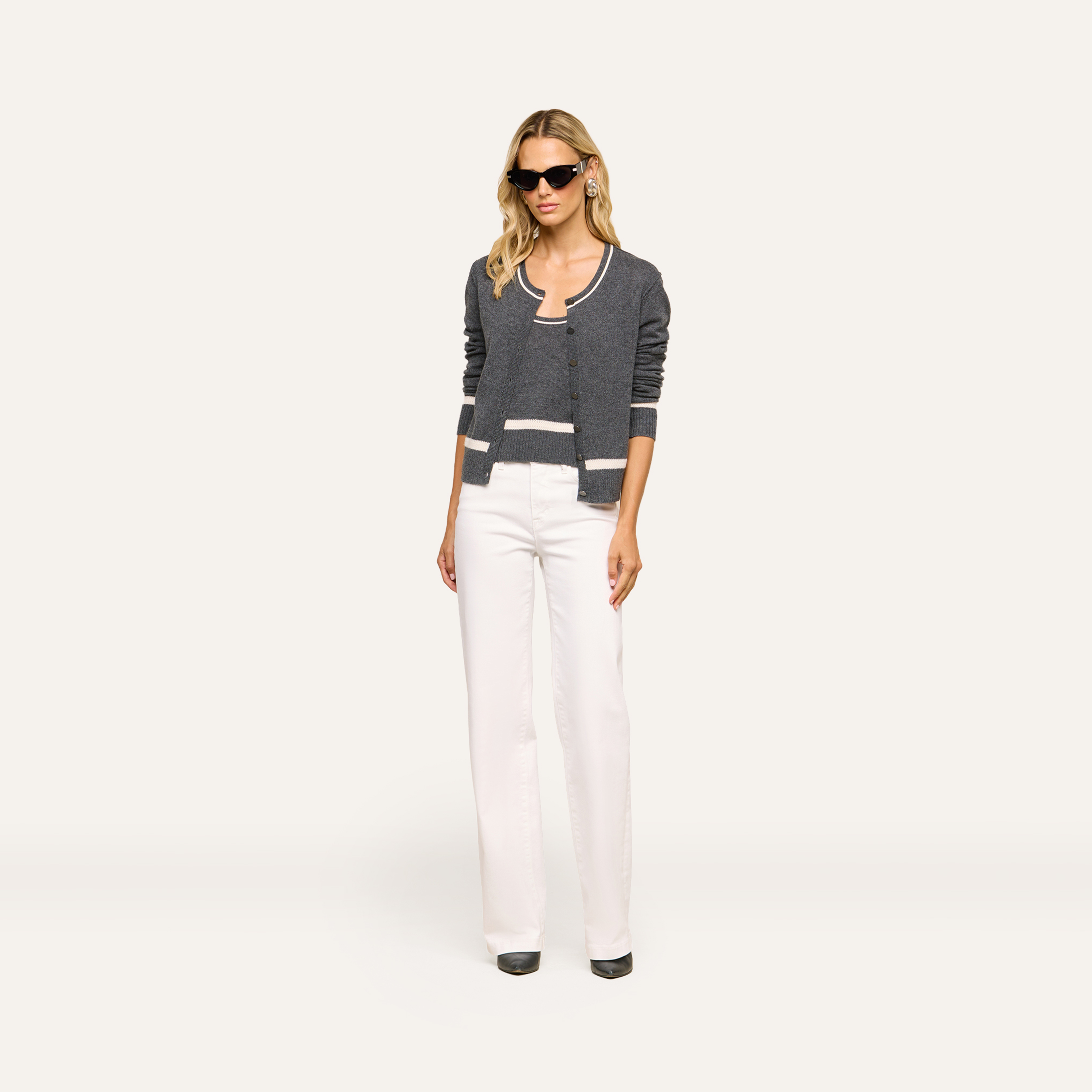 A woman wearing sunglasses, a dark gray cardigan over a matching top with white trim, white high-waisted pants, and black shoes stands confidently against a plain background.