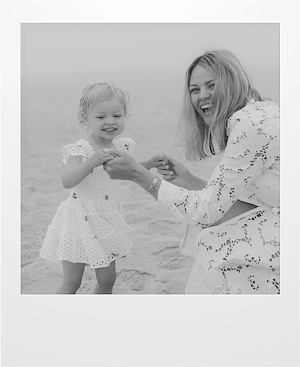 A woman and a young girl, both wearing white lace outfits, smile and hold hands while standing on a sandy beach. The photo is in black and white.