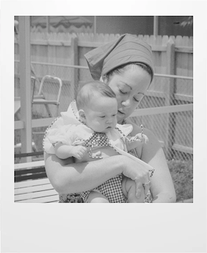 A woman in a headscarf holds a baby in her arms outside near a wooden fence. The baby wears a checkered outfit. Both look down, sharing a quiet moment together.