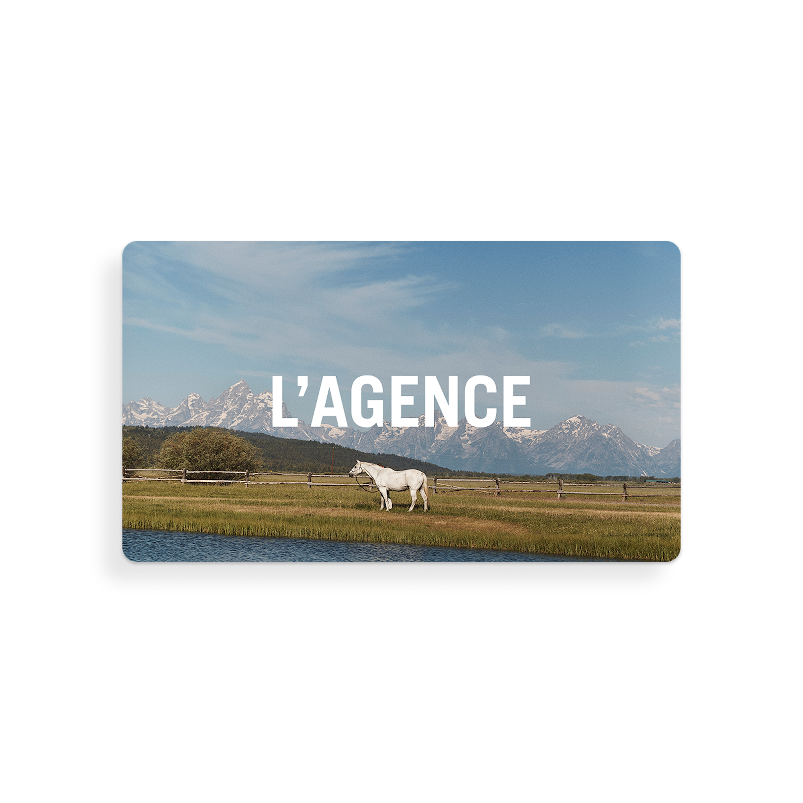 A white horse stands in a grassy field near water, with mountains in the background under a blue sky. The words “L’AGENCE Gift Card” are displayed in large white letters across the sky, evoking the convenient way gift cards and redemption instructions are delivered via email.