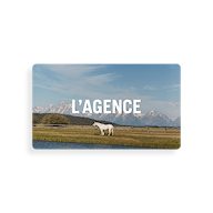 A white horse stands in a grassy field near water, with mountains in the background under a blue sky. The words “L’AGENCE Gift Card” are displayed in large white letters across the sky, evoking the convenient way gift cards and redemption instructions are delivered via email.