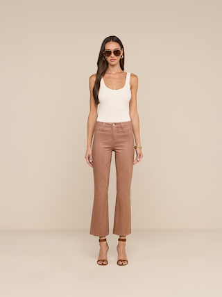 A woman wearing a white tank top, the Petite Kendra Coated Jean in light brown, which is a new Les Petites jean, with heeled sandals, and sunglasses stands against a beige background.
