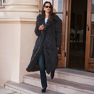 A woman wearing a charcoal-colored shag Lizbeth Faux Fur Jacket, blue jeans, and black pointed-toe boots steps out of a doorway with a relaxed fit.