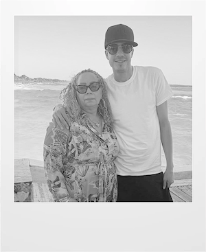 A woman with curly hair and glasses stands beside a man wearing a cap, sunglasses, and a white t-shirt. They are outdoors near the ocean, with waves and a sandy shoreline in the background.