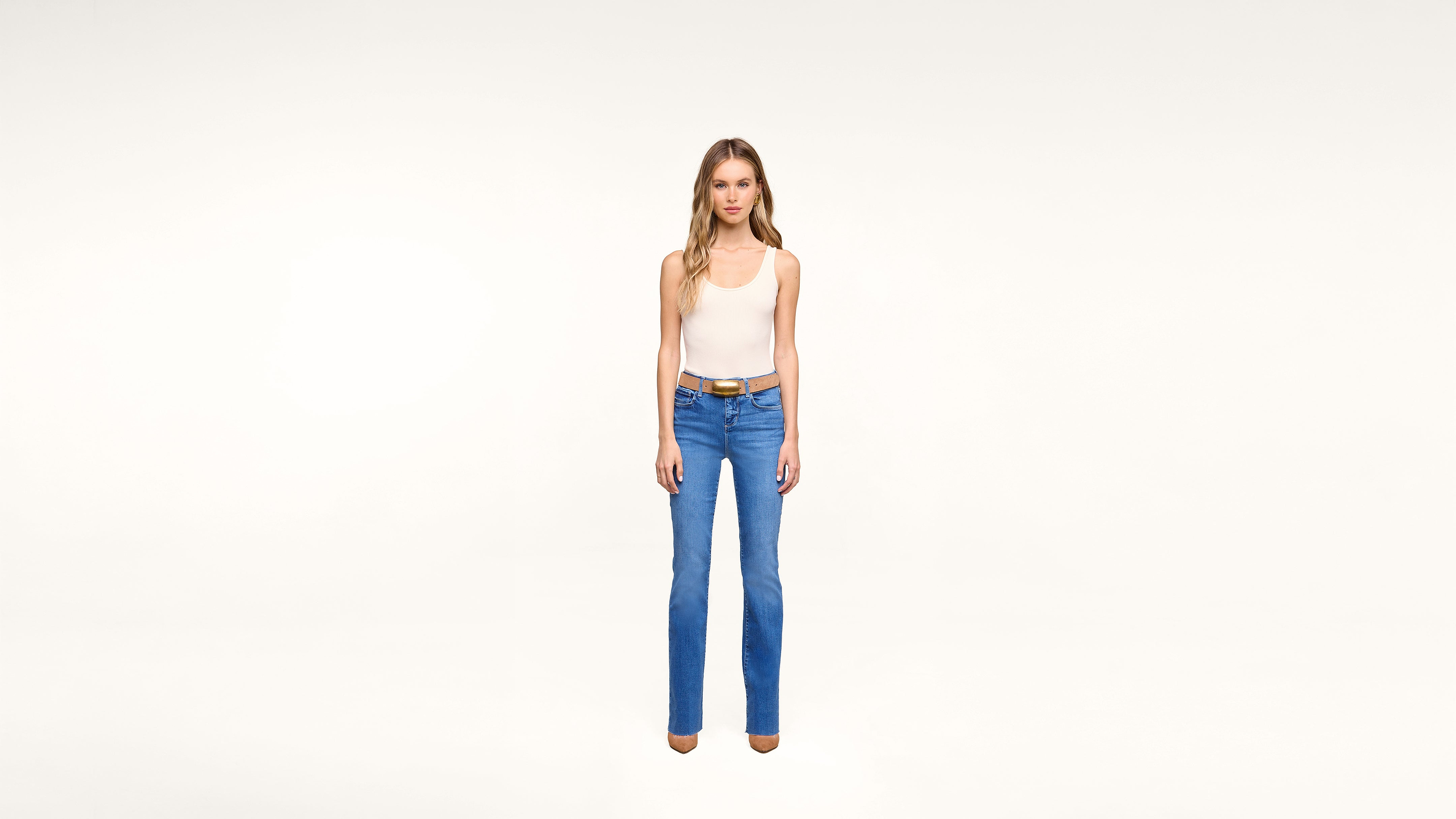 A woman stands against a plain white background wearing a white tank top, blue jeans, a brown belt, and brown shoes, facing forward with a neutral expression.
