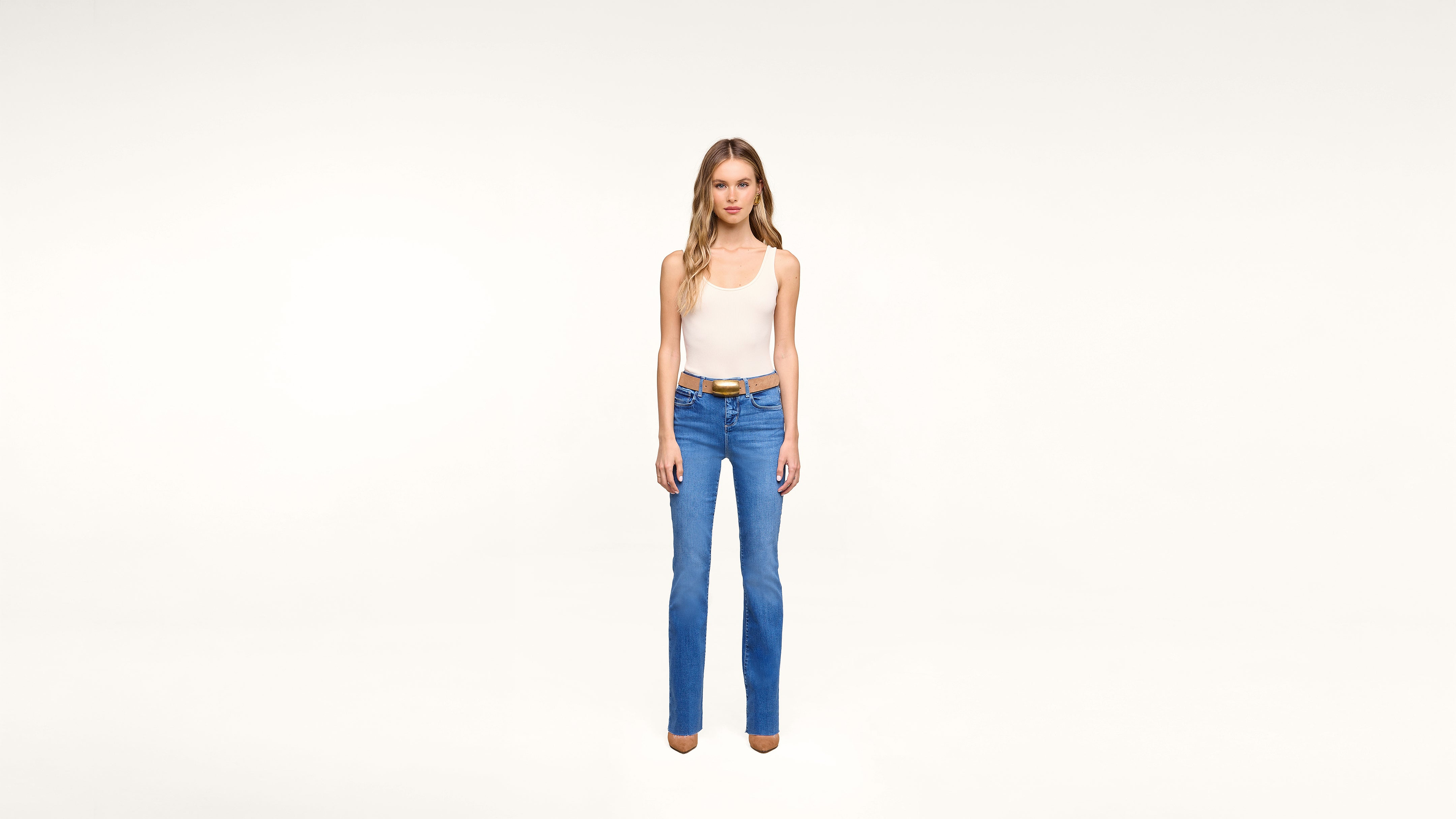 A woman stands against a plain white background wearing a white tank top, blue jeans, a brown belt, and brown shoes, facing forward with a neutral expression.