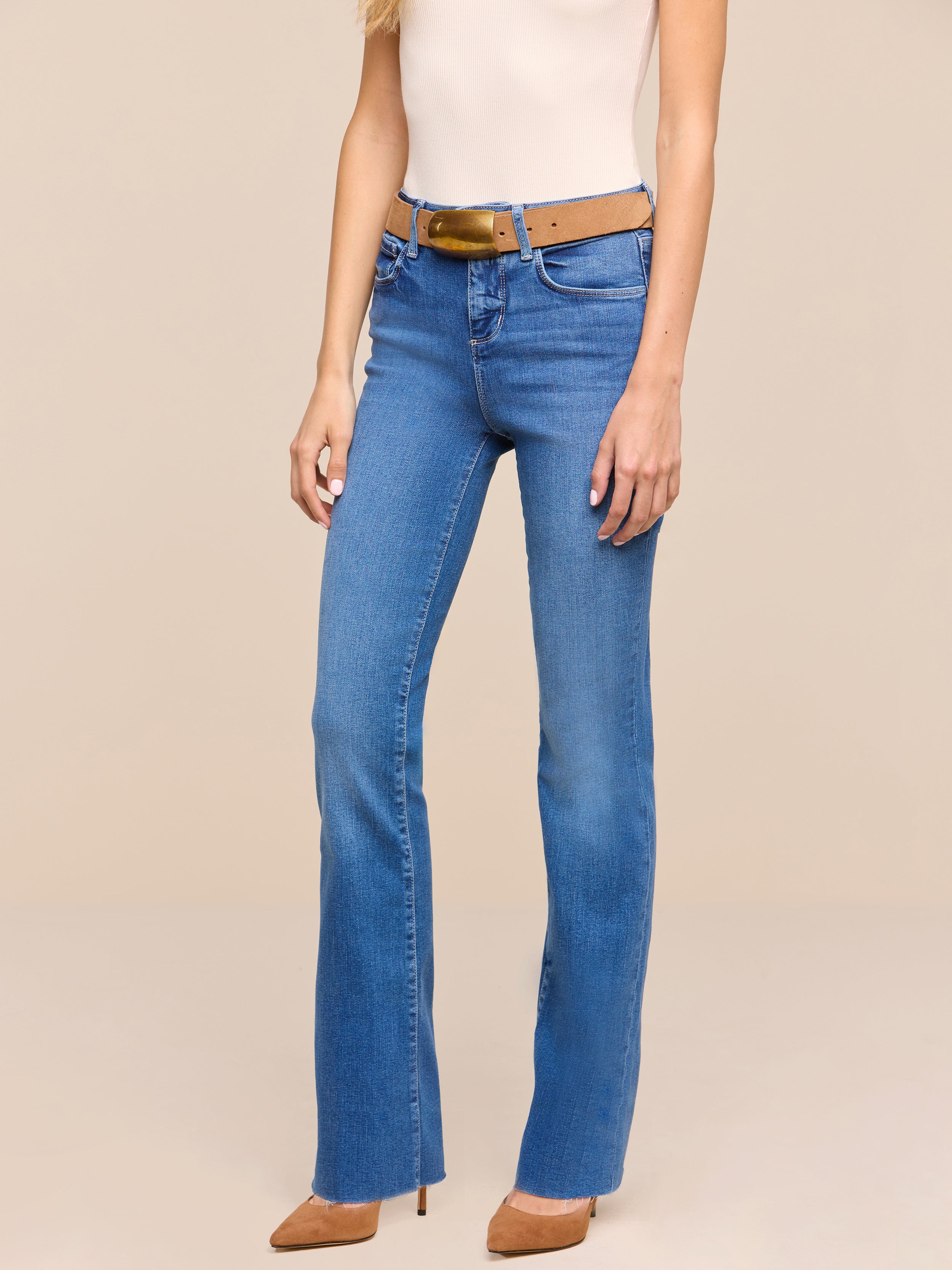 A woman wearing Petite Ruth Straight-Leg Jeans in heritage indigo wash, a tan belt, a sleeveless beige top, and tan high-heeled shoes stands against a plain white background.