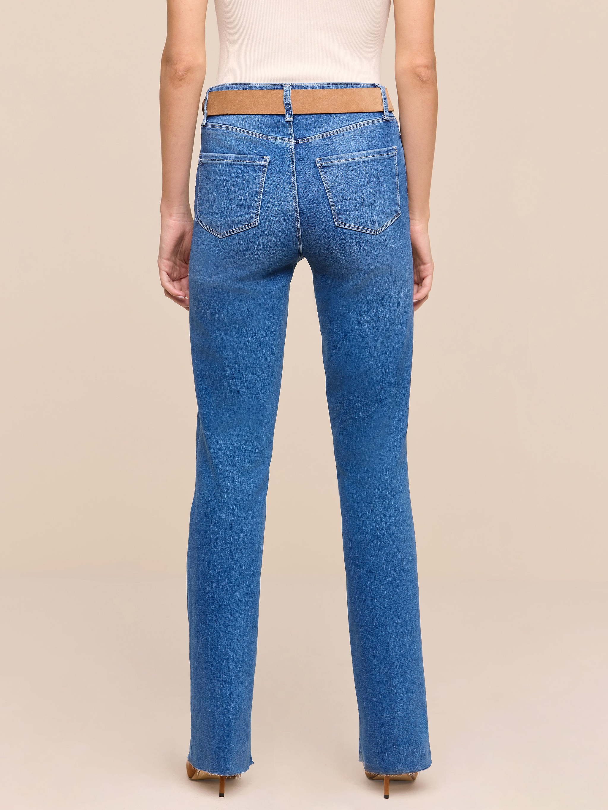 A person stands facing away, wearing the Petite Ruth Straight-Leg Jean in a heritage indigo wash featuring a cut-out section at the waistband, paired with a beige top. The background is plain white.