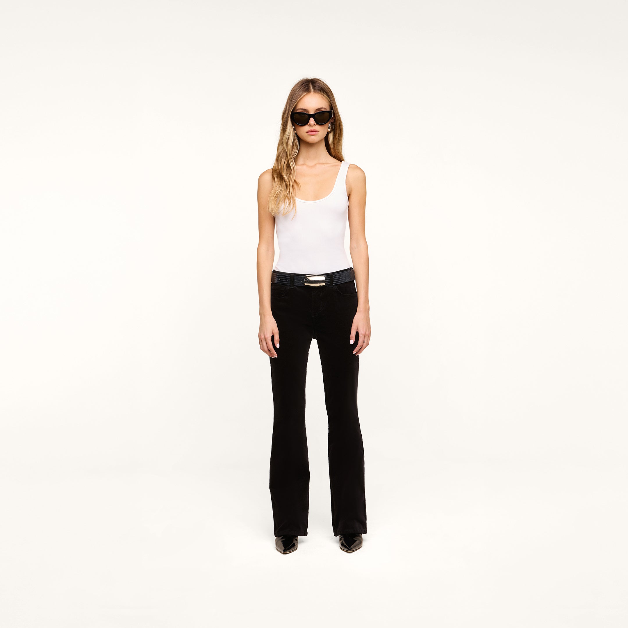 A woman with long hair wearing sunglasses, a white tank top, a black belt, black shoes, and the Petite Stevie Velvet Jeans stands against a plain white background.