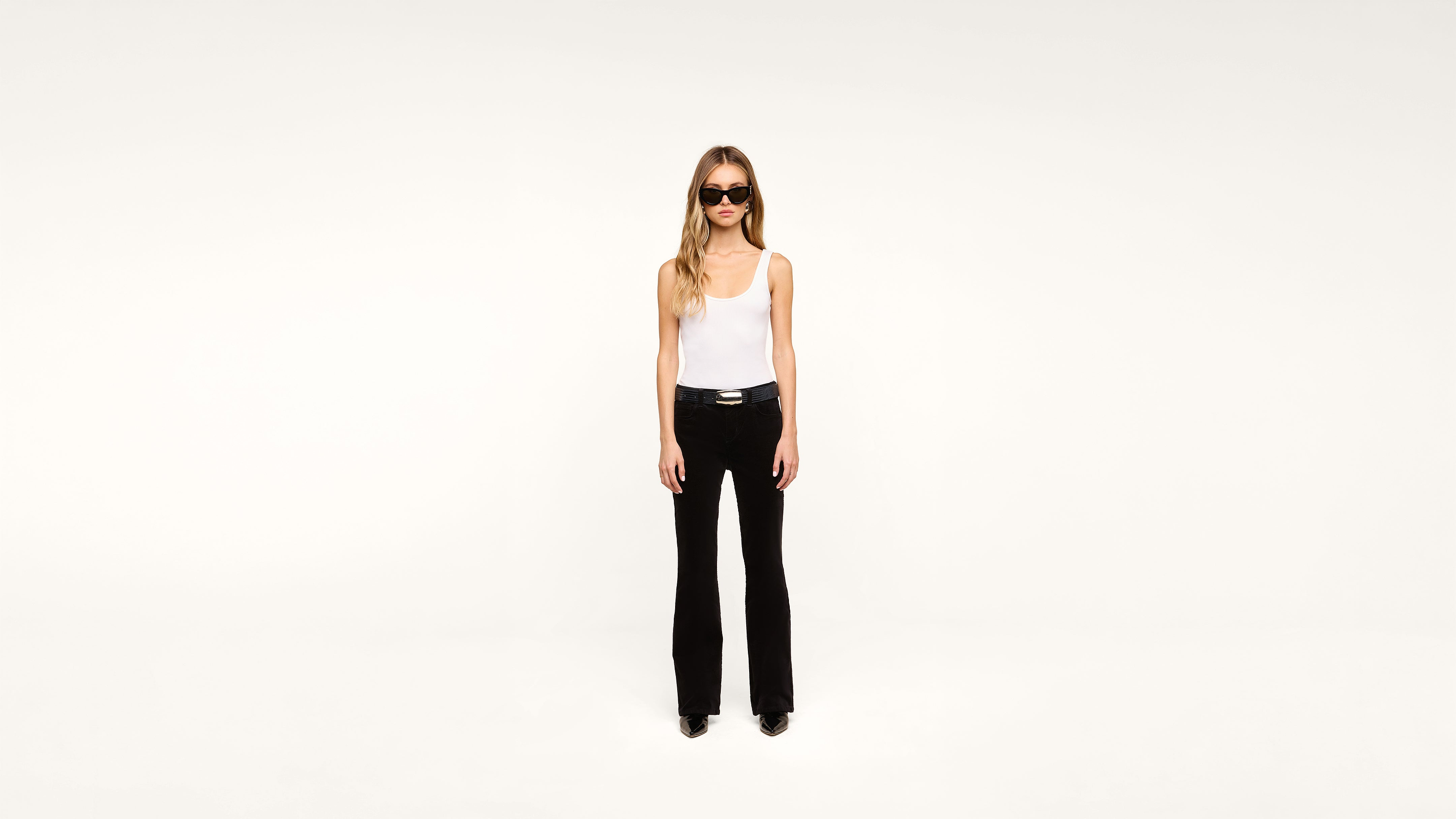 A woman with long hair wearing sunglasses, a white tank top, black pants, and black shoes stands against a plain white background.