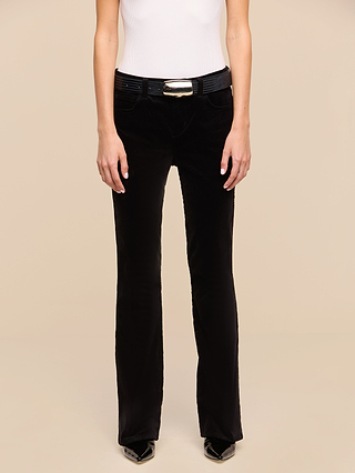 A person wearing a white sleeveless top, black belt with metallic buckle, Petite Stevie Velvet Jeans in a high-rise slim straight-leg fit, and pointed black shoes, standing against a plain beige background.