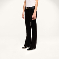 A person wearing the Petite Stevie Velvet Jean with a black belt, a white sleeveless top, and black pointed-toe shoes stands against a plain white background.
