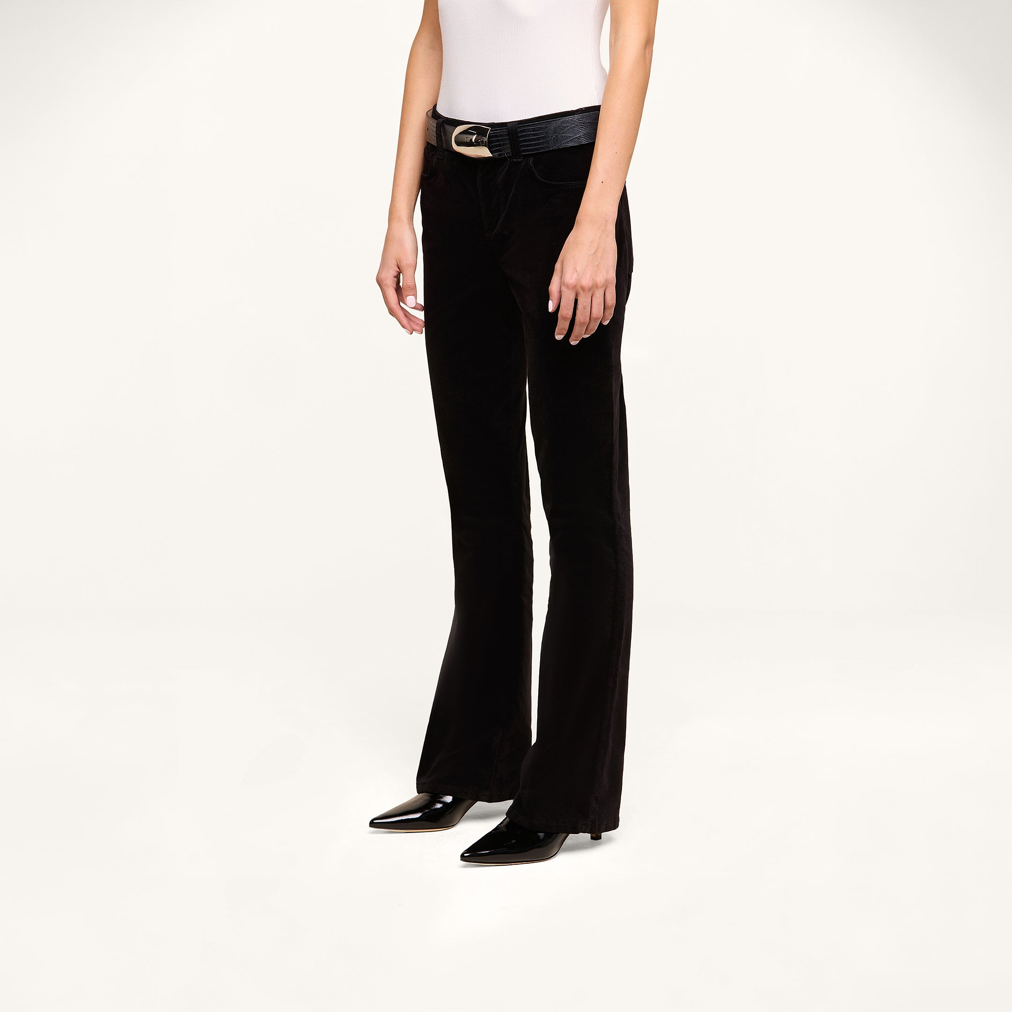 A person wearing the Petite Stevie Velvet Jean with a black belt, a white sleeveless top, and black pointed-toe shoes stands against a plain white background.