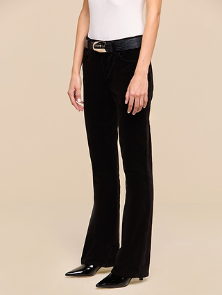 A person wearing a white sleeveless top, the Petite Stevie Velvet Jean in black high rise flared style, a black belt with a gold buckle, and black pointed-toe shoes, standing against a beige background.
