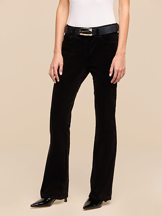 A person is shown from the shoulders down against a beige background, wearing the Petite Stevie Velvet Jean in black with a slim straight-leg fit, paired with a black belt featuring a gold buckle, black pointed-toe shoes, and a sleeveless white top.