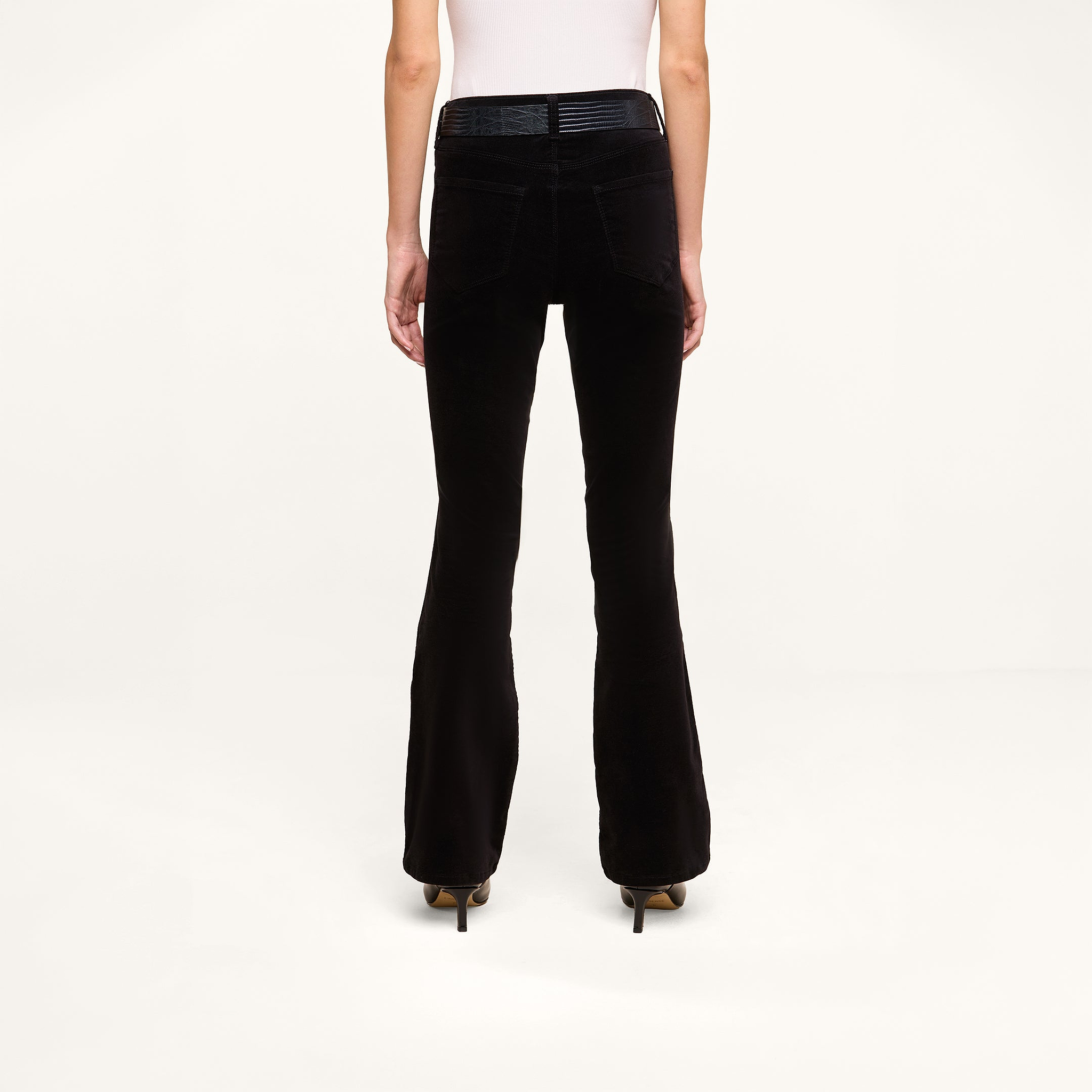 A person is seen from the back, standing against a plain white background, wearing the Petite Stevie Velvet Jean styled with heeled shoes.