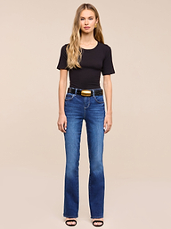 A woman with long blonde hair wears a black t-shirt, the Petite Selma Bootcut Jean in blue super stretch denim, a black belt with a gold buckle, and black sandals, standing against a plain white background.