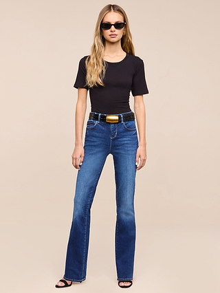 A woman with long blonde hair, wearing sunglasses, a black t-shirt, the Petite Selma Bootcut Jean from the Les Petites Collection in super stretch denim, a black belt with a gold buckle, and black heeled sandals stands against a plain beige background.