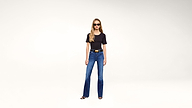 A woman with long blonde hair wearing sunglasses, a black t-shirt, blue jeans, a black belt, and heeled sandals stands against a plain white background.