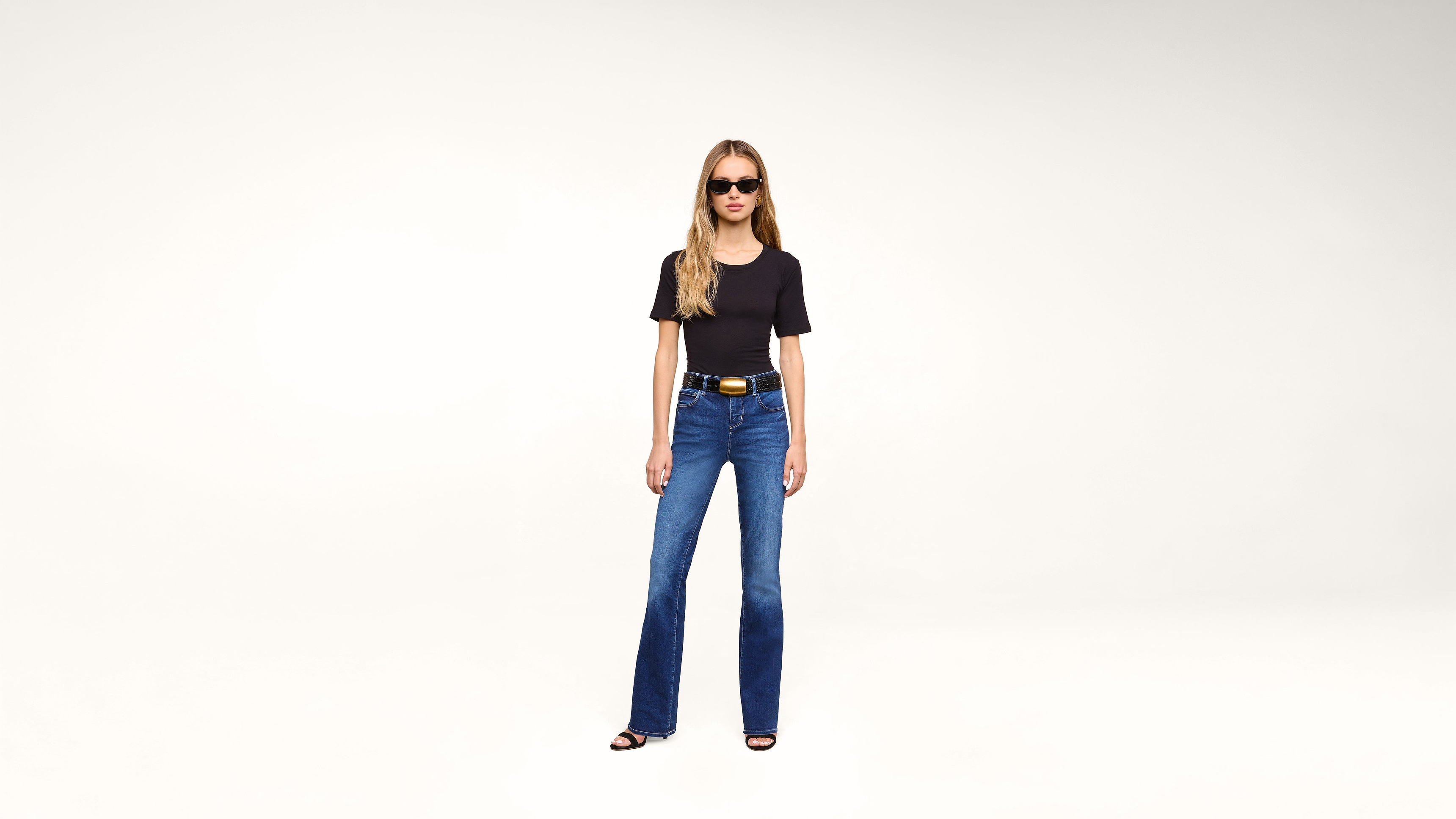 A woman with long blonde hair wearing sunglasses, a black t-shirt, blue jeans, a black belt, and heeled sandals stands against a plain white background.