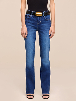 A person wears the Petite Selma Bootcut Jean from the Les Petites Collection in blue, a black top, a black belt with a large gold buckle, and black open-toe heels. The background is plain beige