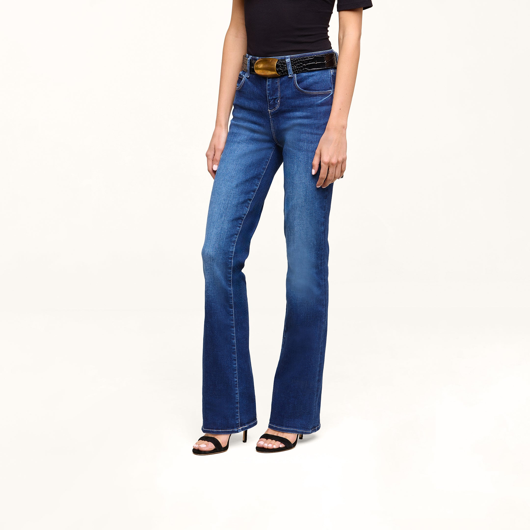 A person wearing a black short-sleeve top, the Petite Selma Bootcut Jean in blue, a black belt, and black high-heeled sandals stands against a plain white background.