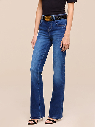 A person wearing the Petite Selma Bootcut Jean from the Les Petites Collection in blue, a black short-sleeve top, a black belt with a large buckle, and black heeled sandals, standing against a beige background.