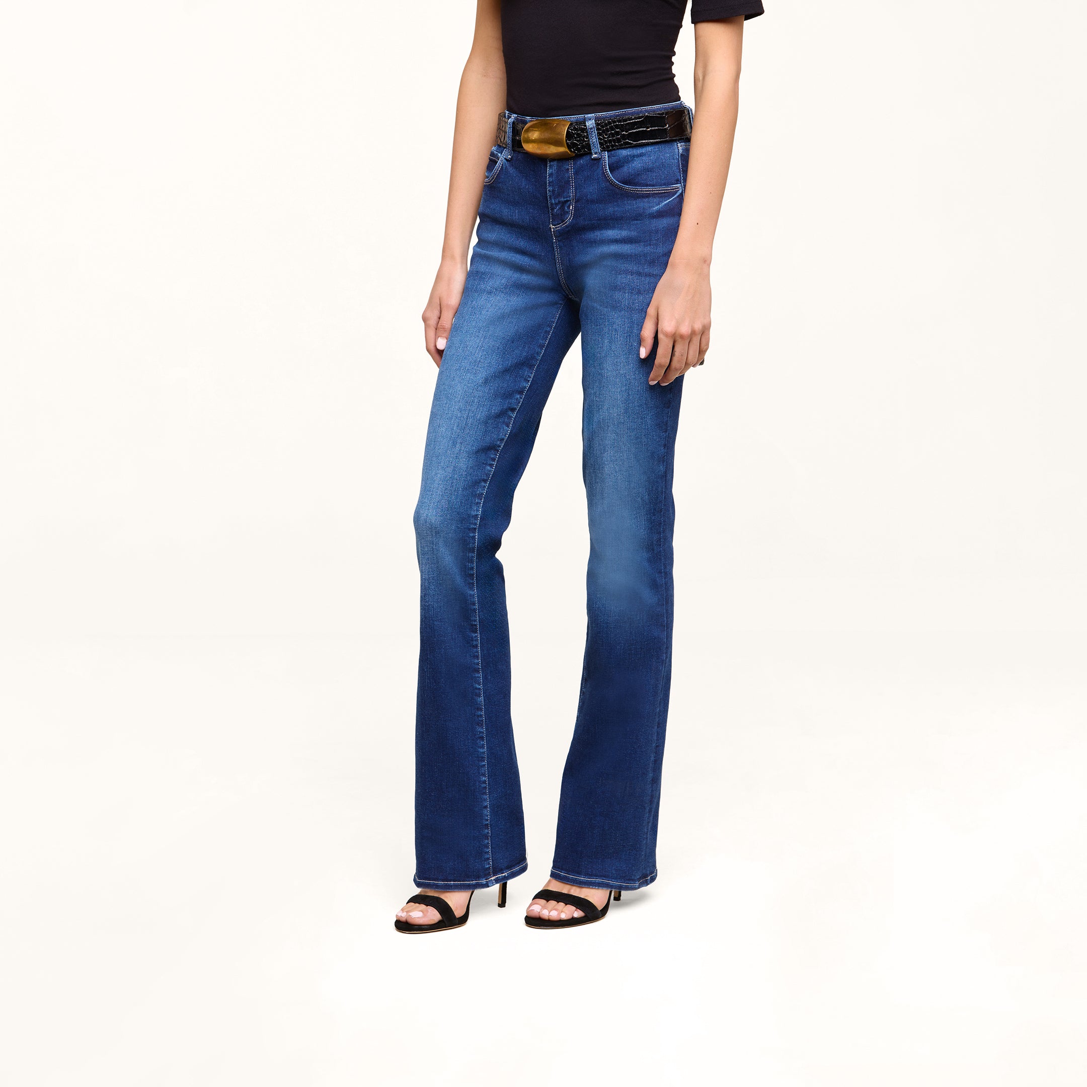 A person wearing a black short-sleeve top, the Petite Selma Bootcut Jean in blue, a black belt, and black high-heeled sandals stands against a plain white background.