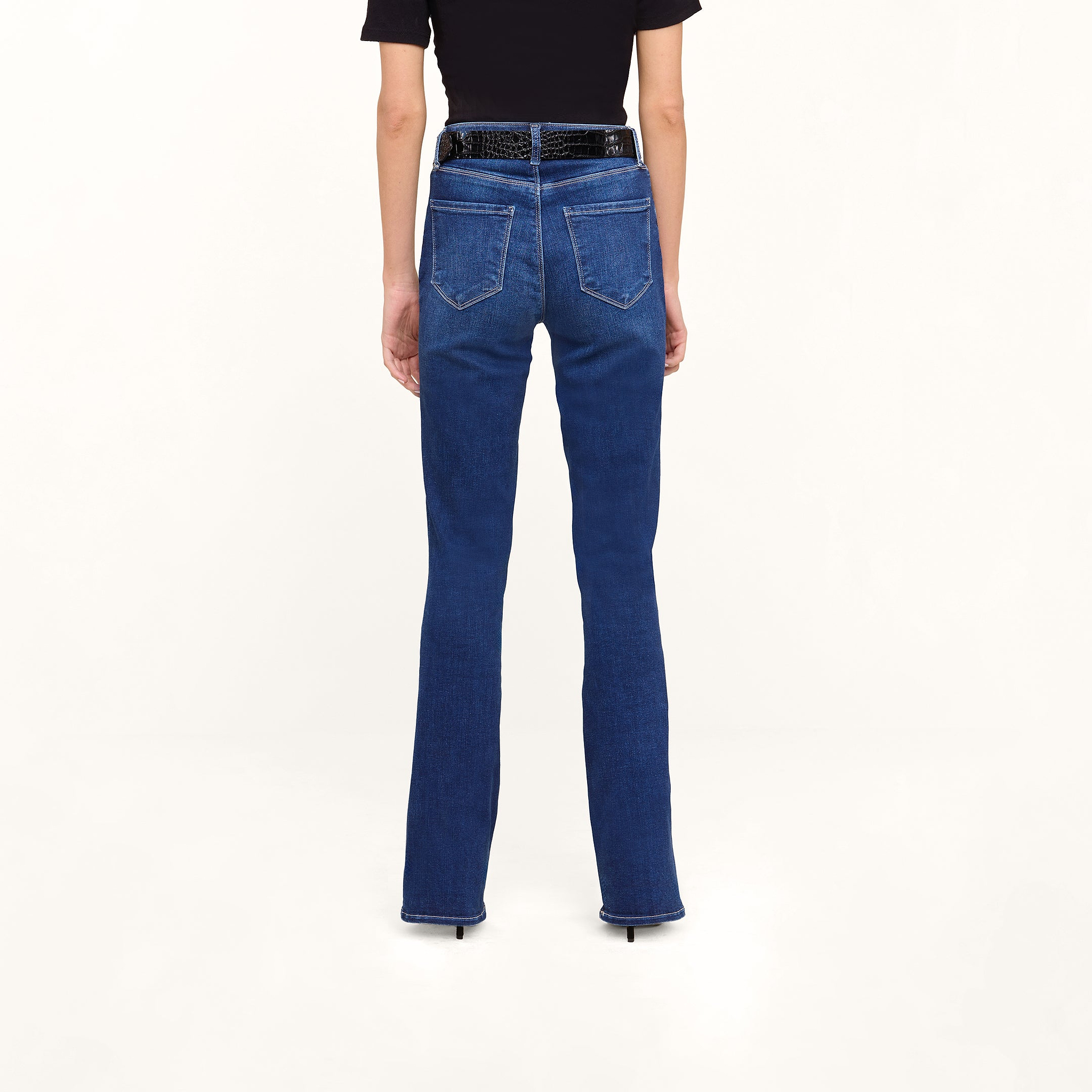 A person stands facing away, wearing Petite Selma Bootcut Jeans in dark blue super stretch denim with a black belt and a short-sleeve black top, against a light background.