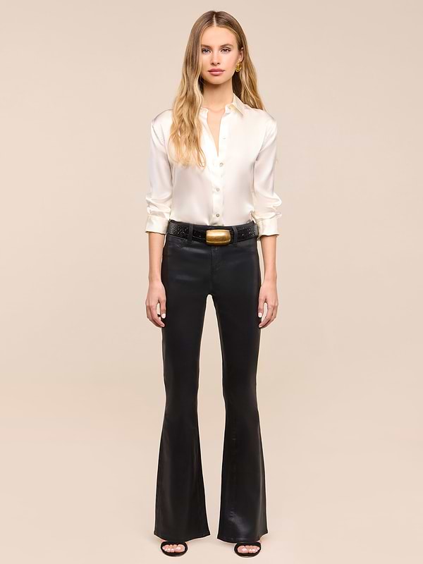 A woman stands against a white background, wearing a white button-up blouse, the Petite Marty Coated Jean in black high-rise petite flare style, a black belt with a gold buckle, and black heeled sandals.
