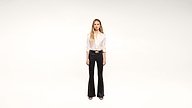 A woman stands against a plain white background, wearing a white blouse, black flared pants, a black belt with a gold buckle, and black shoes.