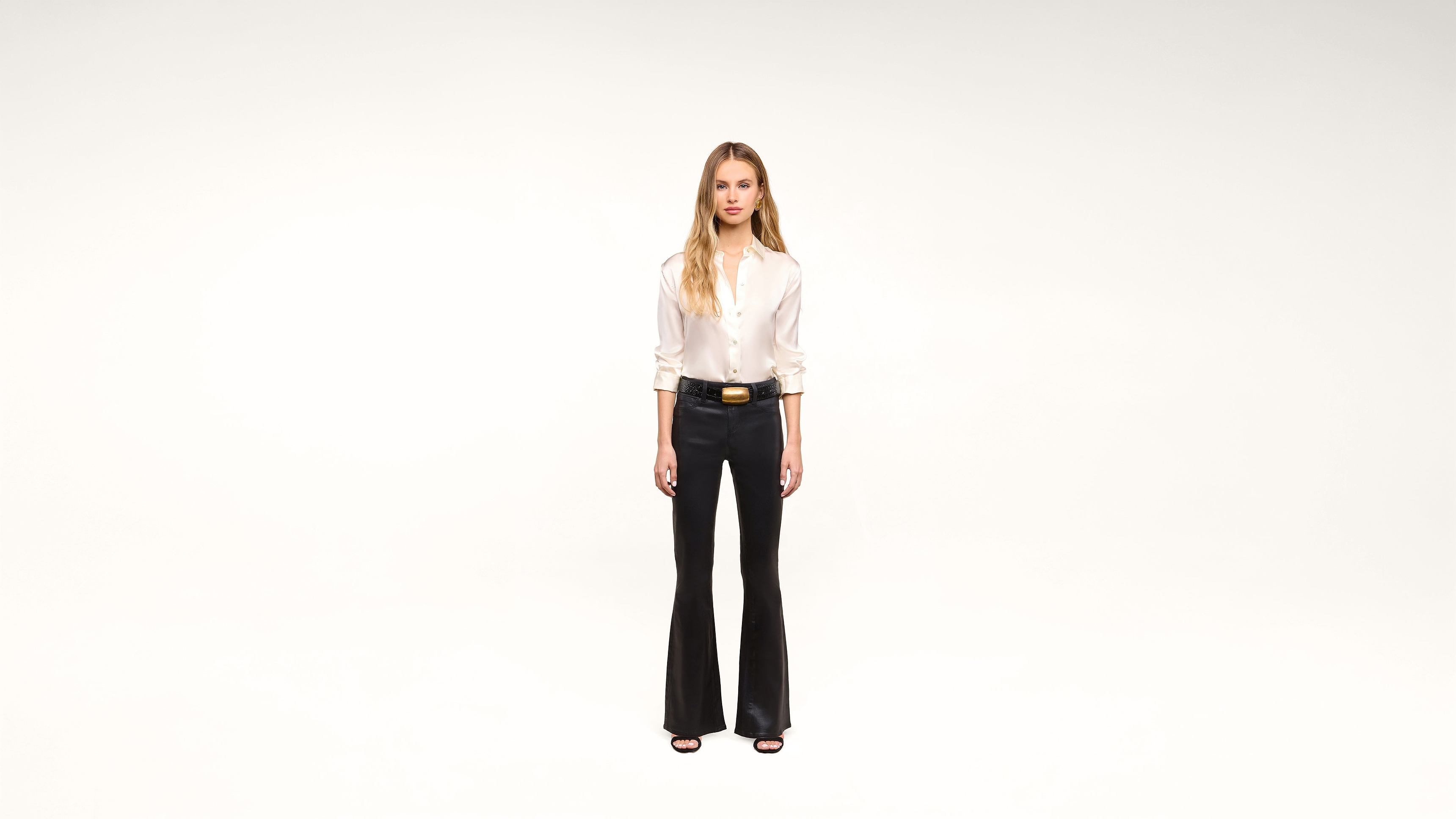 A woman stands against a plain white background, wearing a white blouse, black flared pants, a black belt with a gold buckle, and black shoes.