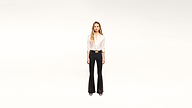 A woman stands facing forward against a plain white background, wearing a stylish white blouse, black flared pants, a chic black belt with a gold buckle, and classic black heels.