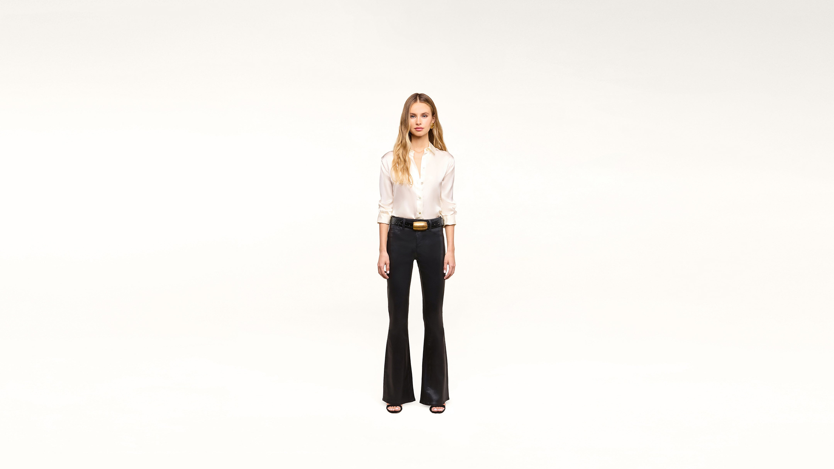 A woman stands facing forward against a plain white background, wearing a stylish white blouse, black flared pants, a chic black belt with a gold buckle, and classic black heels.