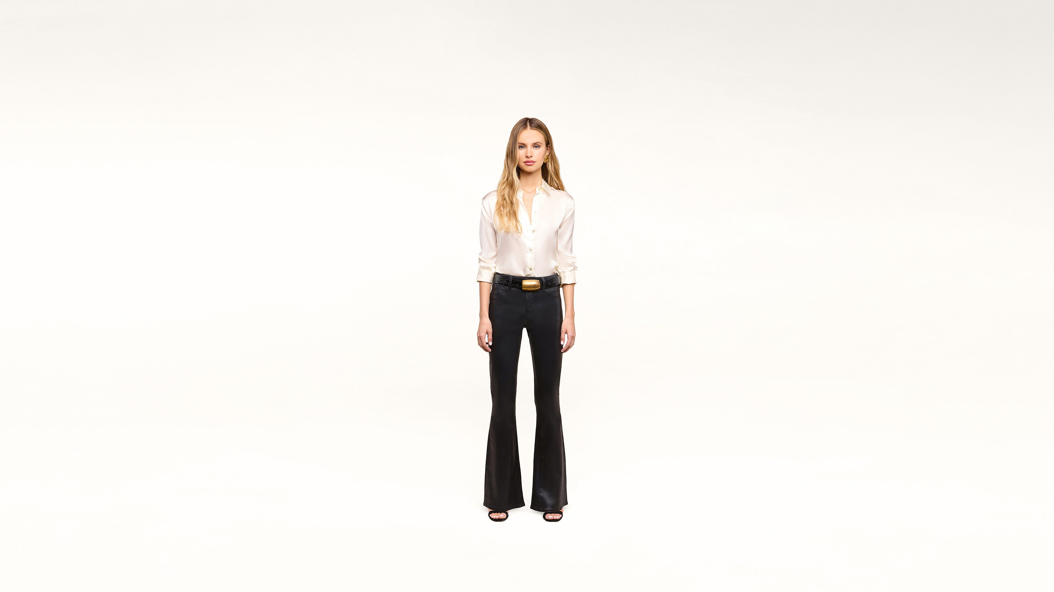 A woman stands facing forward against a plain white background, wearing a stylish white blouse, black flared pants, a chic black belt with a gold buckle, and classic black heels.