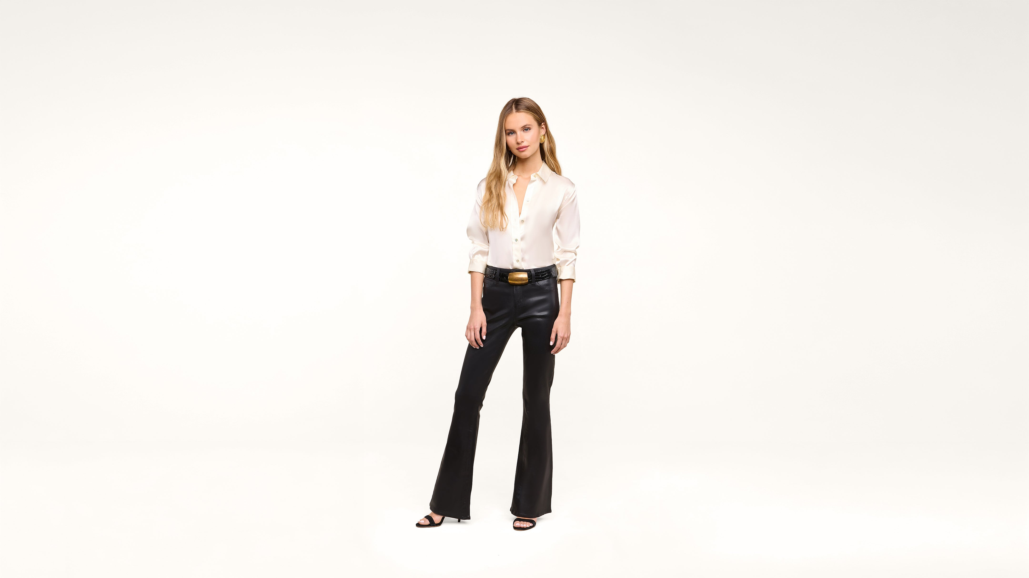 A woman stands in a white studio wearing a white blouse, black leather pants, a black belt with a gold buckle, and black heels. She has long, blonde hair and her hands rest casually by her sides.