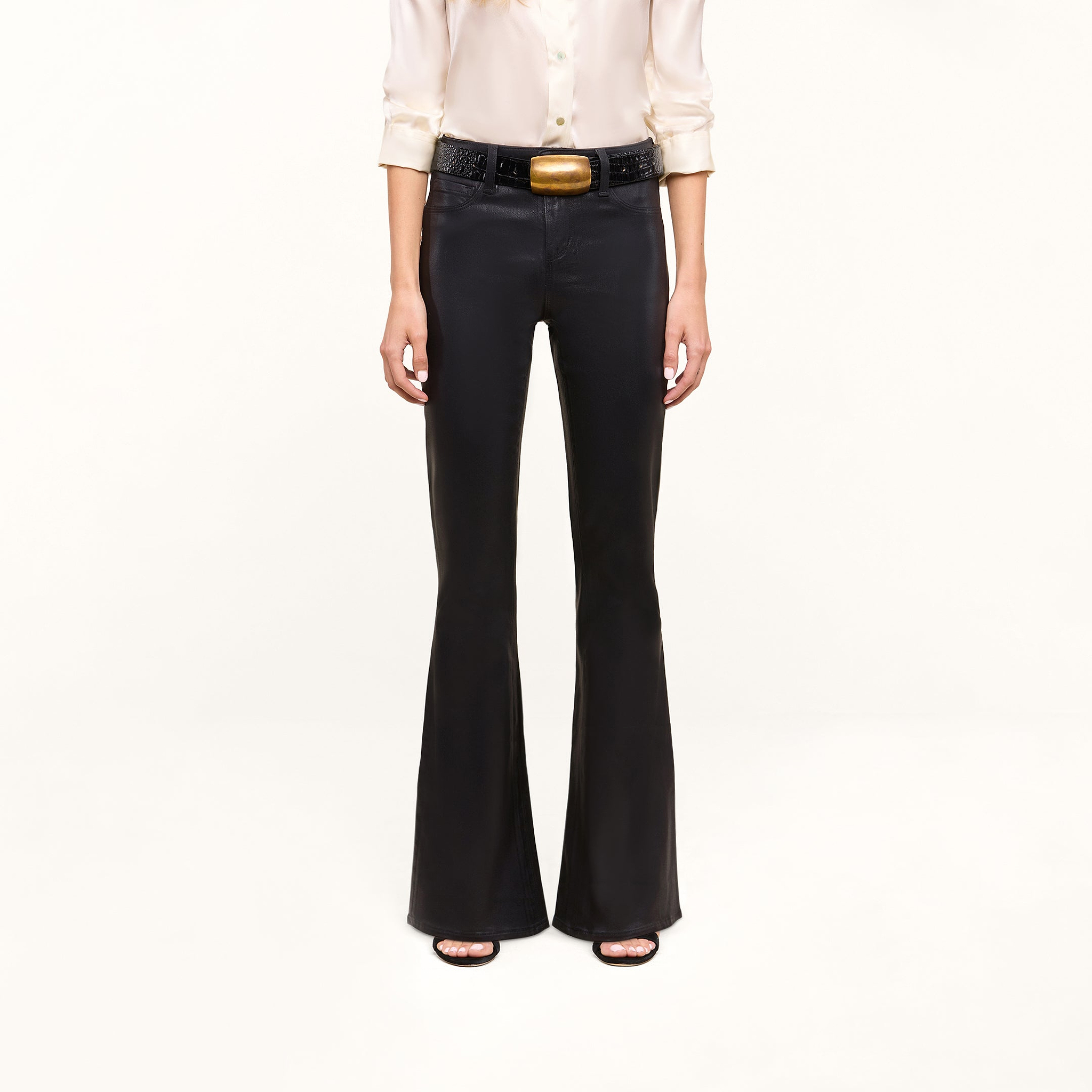 Person wearing a cream blouse, the Petite Marty Coated Jean in black high rise flare style, a black belt with a large gold buckle, and black open-toe heels, standing against a plain light background. Upper face is not visible in the image.