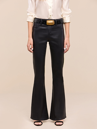 Person wearing a cream satin blouse, Petite Marty Coated Jean from the Les Petites Collection in black high rise petite flare style, a black belt with a large gold buckle, and open-toe black heels, standing against a plain beige background.