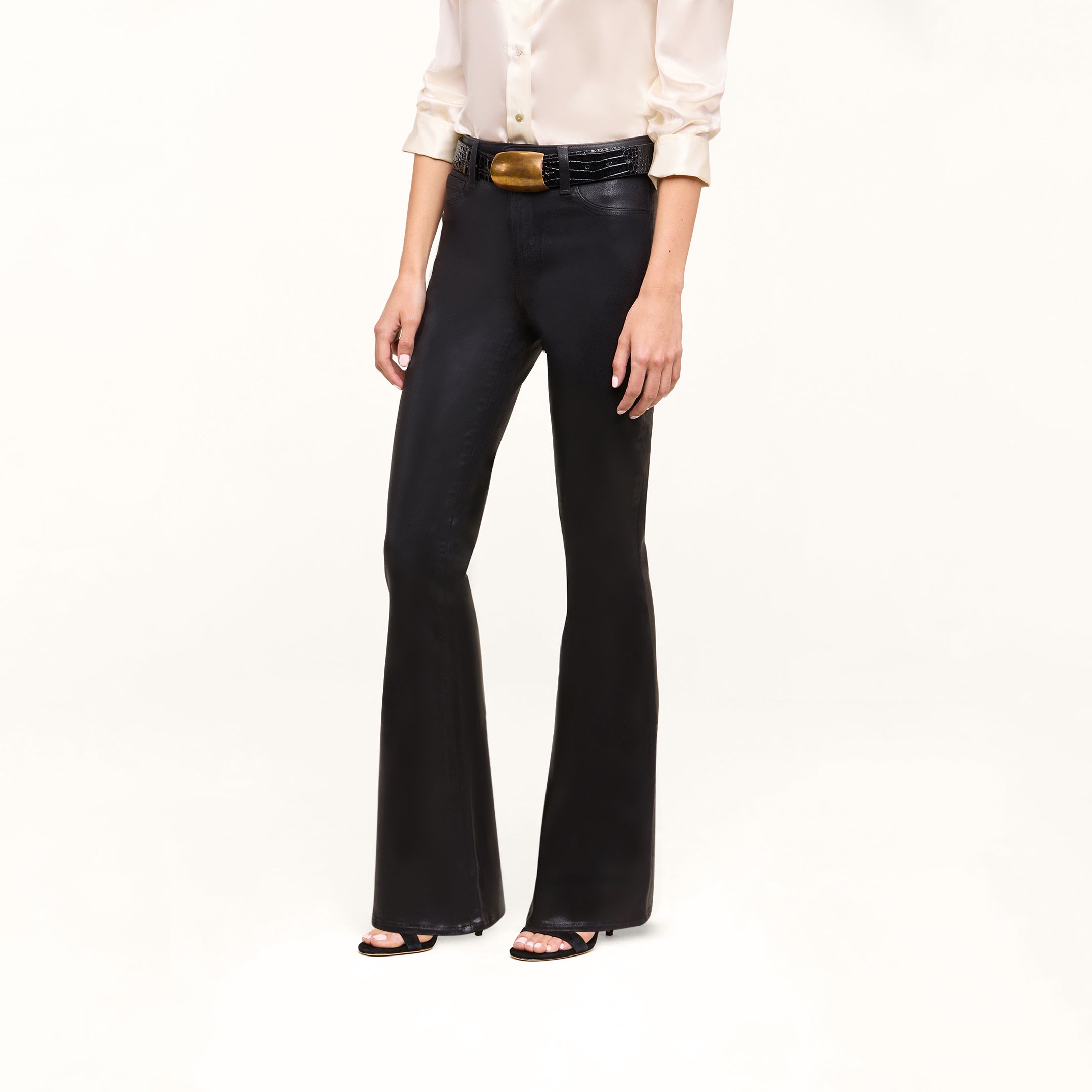 A person wearing a cream blouse, black belt, the Petite Marty Coated Jean in high rise petite flare style, and black heeled sandals stands against a plain white background.