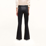 A person stands facing away, wearing the Petite Marty Coated Jean—high-rise black flared pants—paired with a cream long-sleeve shirt tucked in and black high-heeled shoes. The background is plain white.