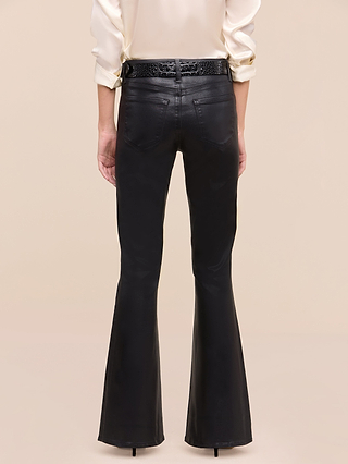 A person is seen from the back, standing against a plain beige background, wearing the Petite Marty Coated Jean in black high rise flared style from the Les Petites Collection, paired with a cream long-sleeve shirt and a black belt.
