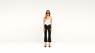 A woman wearing sunglasses, a white tank top, black flared pants, and black heels stands confidently against a plain white background, facing the camera.