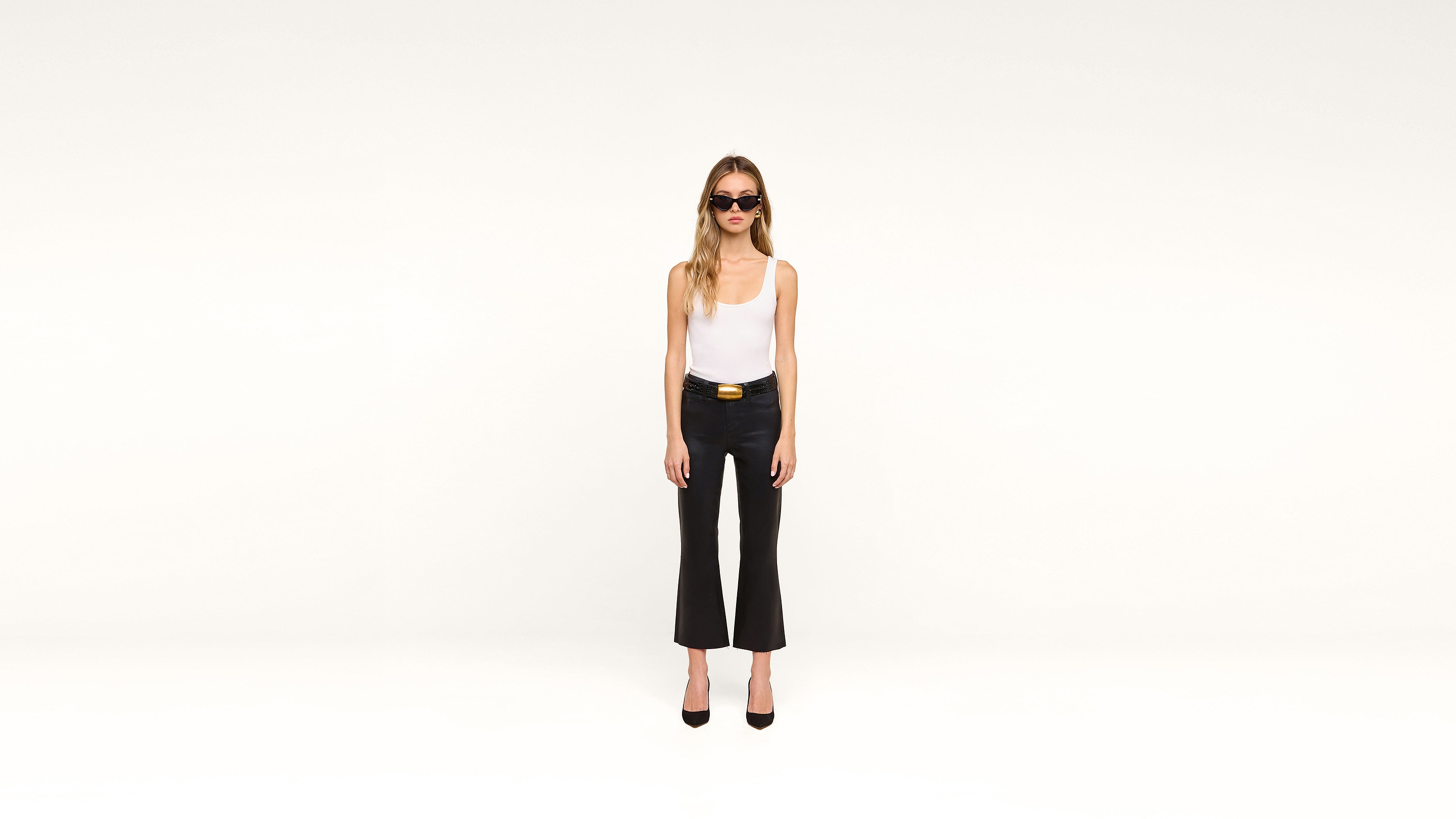A woman wearing sunglasses, a white tank top, black flared pants, and black heels stands confidently against a plain white background, facing the camera.